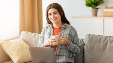 Genç bir kadın rahat bir koltukta oturur, fincandan kahve yudumlar. Önünde dizüstü bilgisayarı açık ve mutlu görünüyor ve öğleden sonra evde işiyle meşgul..