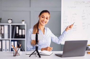 Bir öğretmen rahat bir sınıfta çevrimiçi İngilizce dersi veriyor. Gülümsüyor ve notlarla dolu bir tahtaya işaret ediyor..