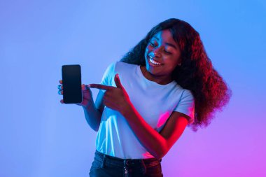 Kıvırcık saçlı genç bir kadın parlak bir stüdyoda akıllı telefonu tutarken gülümsüyor. Arka planda enerjik bir atmosfer yaratan parlak mavi ve pembe ışıklar var..