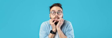 Gözlüklü ve sakallı genç bir adam şok edici bir ifade sergiliyor. Elleri yüzünde ve parlak mavi bir ortamda ürkmüş ya da korkmuş görünüyor..