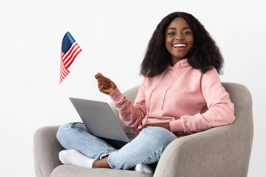 Bir kadın rahat bir sandalyede otururken mutlu bir şekilde gülümsüyor, bir elinde küçük bir Amerikan bayrağı diğer elinde ise dizüstü bilgisayar kullanıyor. Ayar aydınlık ve neşeli, kutlama havasını yansıtıyor..