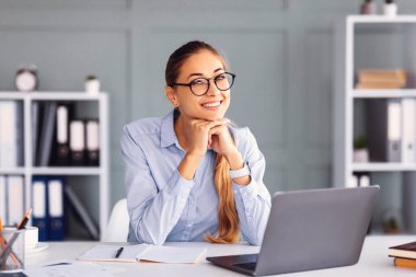 Uzun saçlı bir kadın masasında oturuyor, gülümseyerek çenesini ellerinin üzerinde dinlendiriyor. Etrafında dizüstü bilgisayarı, defterleri ve ofis malzemeleriyle şık bir ofiste..