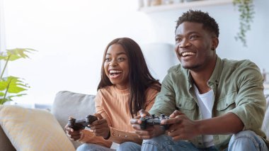 İki arkadaş sıcak bir oturma odasında video oyunu oynuyorlar. Onlar hem gülümseyen hem de ekrana odaklanmış rahat bir kanepede otururlar. Pencereden güneş ışığı akıyor..