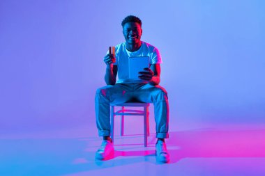 Bir adam bir elinde tablet, diğer elinde kart tutarken gülümseyerek sandalyeye oturur. Sahne parlak mavi ve pembe ışıklarla aydınlatılıyor, canlı bir atmosfer yaratıyor..