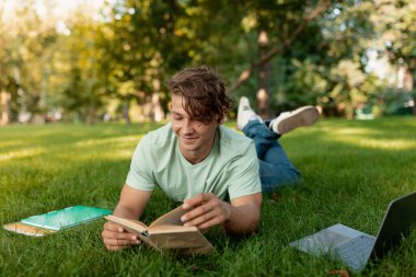 Genç bir adam parkta çimlerin üzerinde uzanmış, kitap okurken gülümsüyor. Yakınlarda not defterleri ve bir dizüstü bilgisayar var. Açık havada ders çalışmanın keyifli bir öğleden sonrası olduğunu gösteriyor..