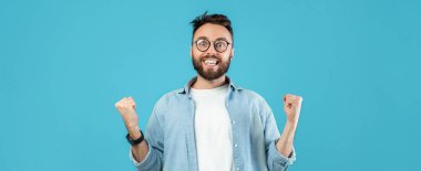 Gözlüklü ve sakallı genç bir adam yumruklarını havaya kaldırarak neşesini gösteriyor. Yüzünde kocaman bir gülümseme var, mutluluk saçıyor. Parlak mavi arka plan neşeli havayı arttırır..