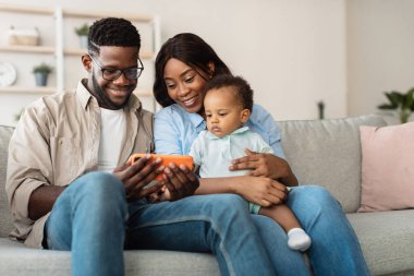 Genç Afrikalı Amerikalı ebeveynler küçük oğullarıyla kanepede dinleniyorlar. Birlikte eğlenceli bir eğitim uygulaması keşfediyorlar. Eğlenceli bir aile bağı kuruyorlar..