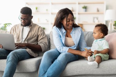 Parlak bir oturma odasında, siyah bir anne kanepeye oturur ve bebeğiyle mutlu bir şekilde oynar. Bu arada, baba dizüstü bilgisayarına odaklanır, evde iş ve aile zamanını dengeler..