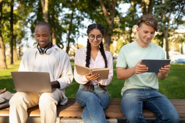 Three young adults sit on a bench in a park, each focused on their devices. One uses a laptop, another reads a notebook, and the last browses on a tablet under clear skies.
