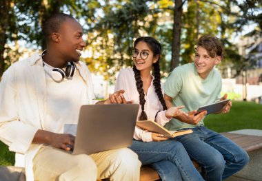 Three friends are sitting on a bench in a park, smiling and discussing while using a laptop and tablets. The scene captures the essence of friendly collaboration and learning.