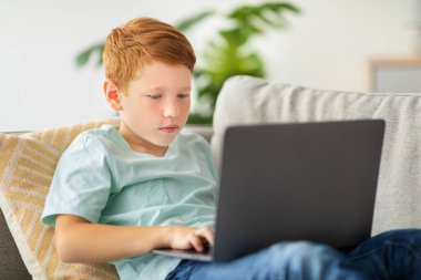 A cheerful redhead boy is reclining on a couch with his laptop, fully engaged in an online video game. He looks happy while enjoying his time alone at home in a modern living room.