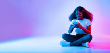 Kıvırcık saçlı genç bir kadın düz bir zeminde bacak bacak üstüne atarak oturur. Etrafı canlı mor ve pembe ışıklarla çevrili akıllı telefonuyla enerji dolu bir atmosfer yaratıyor..