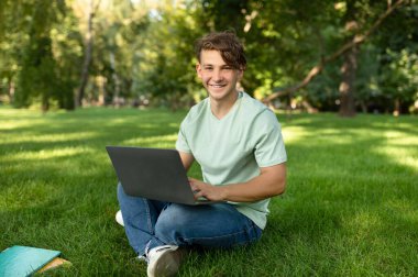 Genç bir adam parktaki çimlerin üzerinde oturuyor, dizüstü bilgisayarına odaklanmış. Güneş ışığı ağaçların arasından süzülür ve işine odaklanırken mutlu görünür..
