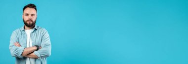 Uzun sakallı genç bir adam canlı mavi bir duvarın karşısında dururken kollarını çapraz yapar. Ciddi bir ifadesi var ve açık mavi bir tişört ve beyaz bir tişört giyiyor..