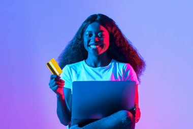 Neşeli genç bir kadın kucağında dizüstü bilgisayarla, elinde kredi kartıyla rahatça oturur. Sahne canlı renklerle aydınlatılıyor, canlı bir atmosfer yaratıyor..