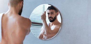 Bir adam, yuvarlak bir aynanın önünde görünüşünü düzeltmeye odaklanır. Ayar parlak bir banyo ve pencereden içeri ışık giriyor..