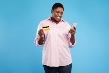 Kısa saçlı bir kadın akıllı telefonuna bakarken gülümsüyor. Elinde bir kredi kartı tutuyor. Arkaplan parlak mavi bir renk, enerjik bir atmosfer yaratıyor..