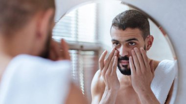 Bir adam banyo aynasının önünde duruyor ve yüzüne cilt bakımı ürünleri uyguluyor. Sabah ışığı pencereden süzülerek rutinine odaklanırken sakin bir atmosfer yaratıyor..