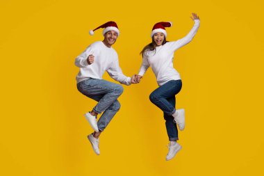 Mutlu bir çift, Noel Baba şapkaları ve günlük kışlık kıyafetlerle el ele tutuşurken heyecanla zıplıyor. Parlak gülümsemeleri ve parlak sarı arkaplanlarıyla neşeli bir tatil ruhu yaratırlar..