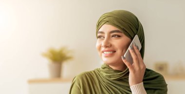 Yeşil başörtüsü takan bir kadın kapalı bir alanda telefonuyla konuşurken gülümsüyor. Atmosfer sıcak ve davetkar görünüyor. Arka planda yeşillik var..
