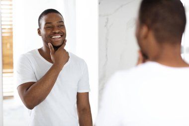 Parlak bir banyoda bir adam aynanın karşısında duruyor, gülümsüyor ve tımar ederken çenesine dokunuyor. Doğal ışık modern ortamı ve kendine güveni arttırır..