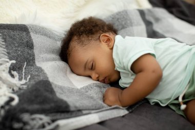 Yakından bakınca, sevimli bir Afro-Amerikan bebeğin rahat bir yatakta yan yattığını görüyoruz. Yumuşak bir battaniyeye sarılmış ve sıcak bir atmosferde huzurlu bir günün tadını çıkarıyor..