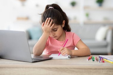 Bir kız dizüstü bilgisayarla masada oturuyor, deftere yazarken stresli görünüyor. Alnını tek eliyle tutuyor, renkli kalemler ve kalemlerle çevrili..