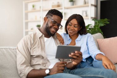 Mutlu bir Afro-Amerikan çifti koltukta rahatça oturup, interneti ve mağazayı gezmek için tablet kullanıyor. Aynı gülümsemeyi paylaşıyorlar ve birbirlerinin gözlerinin içine bakıyorlar. Birlikte güzel vakit geçiriyorlar..