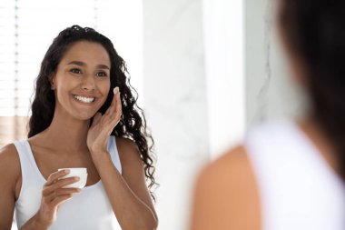 Kıvırcık saçlı bir kadın modern bir banyoda cilt bakımını sever. Yüzüne krem sürüyor. Yansımasına bakıp gülümsüyor, kendine bakmasını teşvik ediyor..