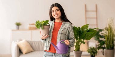 Bir kadın, elinde bir saksı çiçeği ve sulama tenekesi tutarken gülümsüyor, modern bir oturma odasında yemyeşil ile çevrili. Parlak ve davetkar, burası onun bahçe işlerine olan tutkusunu gösteriyor..