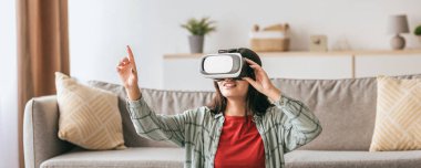 Sanal gerçeklik kulaklığı takan bir kadın oturma odasında ilginç bir deneyimden zevk alır. Mutlu görünüyor ve etrafındaki sanal çevreyle aktif olarak etkileşime geçiyor..