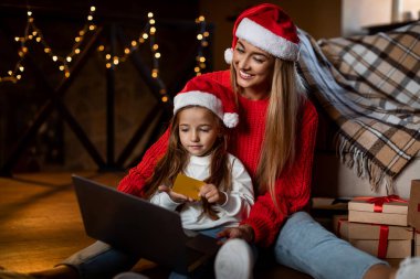 Bir anne ve küçük kızı Noel Baba şapkaları ve rahat kazaklar giyerek yerde oturuyorlar. Online alışveriş yaparken önlerinde bir dizüstü bilgisayar ile neşeli bir anı paylaşıyorlar..