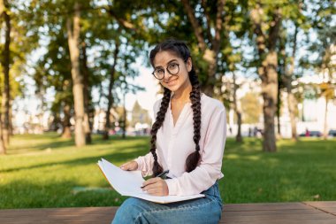 Genç bir kadın parktaki bir bankta rahat bir şekilde oturur, büyük bir kağıt üzerinde bir kalemle çizim yapar. Ağaçlarla çevrili güneşli bir öğleden sonranın tadını çıkarıyor..
