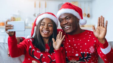 Noel süveteri ve kırmızı Noel Baba şapkası giymiş neşeli bir çift bir koltuğa oturur. Gülümsüyorlar ve selfie çekerken kameraya el sallıyorlar. Oda tatil için sıcak bir şekilde dekore edildi..