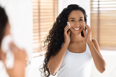 Kıvırcık saçlı bir kadın yüzüne cilt bakım ürünü sürerken gülümsüyor. Parlak bir odada panjurlarla duruyor, güzellik rutininin tadını çıkarıyor..