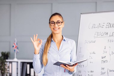 Kendine güvenen bir öğretmen modern bir sınıfta elinde bir defter tutarken baş parmak hareketini gösterir..