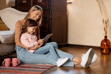 A mother is sitting on the floor with her young daughter, both smiling as they look at a tablet together. They are surrounded by a warm and inviting living room, creating a sense of family joy.