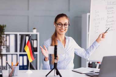 Bir kadın şık bir ofisten internet üzerinden Almanca dersi veriyor. Gülümsüyor, gözlük takıyor ve masasını küçük bir Alman bayrağı süslerken beyaz tahta kullanıyor..