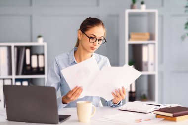 Gözlüklü odaklanmış bir kadın masasında kağıtları inceliyor. Etrafı dizüstü bilgisayar, kahve fincanı ve organizasyonel malzemelerle çevrili. Çalışma alanı parlak ve düzenli..