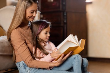A mother sits on the floor, holding a book and reading to her young daughter. They share a warm moment surrounded by a cozy, welcoming living room.