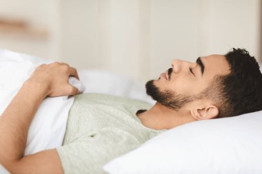 Genç bir adam rahat yatak odasında huzurlu bir uyku çeker. Yumuşak bir battaniyenin altına uzanır ve elleri göğsünde dinlenir. Huzur dolu bir an yaşar..