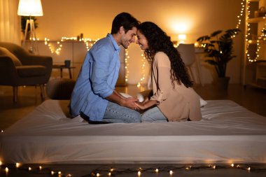 Sevgi dolu bir çift sıcak bir yatak odasında bir yatakta otururken el ele tutuşup alınlarına dokunur. Romantik bir gece buluşmasında duygularını ifade ederler, yumuşak ışıklarla çevrili..