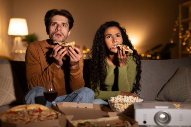 Shocked spouses sit on the couch in their living room, watching a horror film while eating pizza. The cozy atmosphere highlights their family movie night on a weekend.