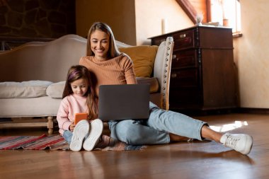A mother sits on the floor with her daughter, both engaged with their devices. The cozy living room features a sofa and warm lighting, creating a pleasant atmosphere for connection.