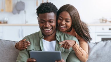 İki insan bir kanepeye yakın oturuyor, birlikte tablete bakıyor ve gülümsüyor. Mutlu görünüyorlar ve bu işle meşguller. Ayar parlak ve davetkar bir yaşam alanı..