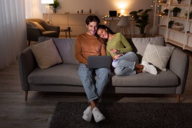 Mutlu çift dizüstü bilgisayarlarında film izlerken koltukta dinleniyor. Evlerinde rahat bir akşam geçiriyorlar, birbirlerine eşlik ediyorlar ve internette geziniyorlar, sıcak bir ortam yaratıyorlar..