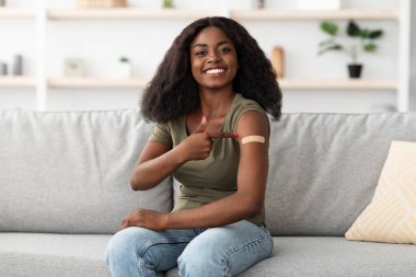 Genç bir kadın aşı olduktan sonra gülümser ve kolunu bandajla gösterir. Parlak oturma odasındaki gri bir kanepede rahatça oturuyor..