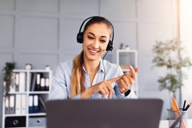 Kulaklık takan bir kadın video görüşmesi yaparken gülümsüyor. Elleriyle hareket ediyor ve masasında oturuyor. Parlak ve düzenli bir ofis alanında..