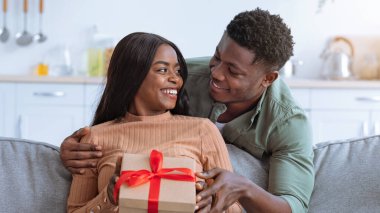 Bir çift, güzel paketlenmiş bir hediyeyi sunarken hassas bir anın tadını çıkarır. Konforlu oturma odası onların neşeli ifadelerini ve sıcak bağlantılarını arttırır..