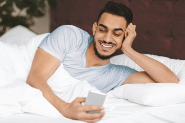 A young man is lying in bed, wrapped in white blankets. He has a relaxed posture and a smile as he looks at his smartphone, enjoying his morning routine.